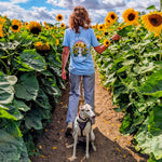 A woman stands in a sunflower field with a dog, viewed from behind. She is wearing a light blue graphic T-shirt that reads "life is better with my dog"