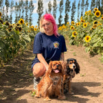 A woman with bright pink hair kneels in a sunflower field with two dogs in front of her. She is wearing a navy blue t-shirt with a colorful graphic on the chest that reads "life is better with my dogs"