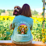 A woman and her dog sit on a wooden bench in a sunflower field, viewed from behind. The woman wears a light blue graphic T-shirt with the text 'LIFE IS BETTER with my dog' and a dog-and sunflower design on the back.