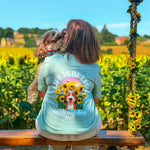 A woman sits on a wooden swing in a sunflower field, seen from behind, wearing a light green graphic T-shirt with a dog and sunflower design. A small dog is on her shoulder, and another dog is on the ground nearby.