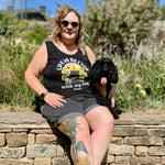 A woman sitting outdoors on a stone wall next to a small black dog, wearing a black graphic vest top that reads 'I'm Better with my Dog' and dark shorts.