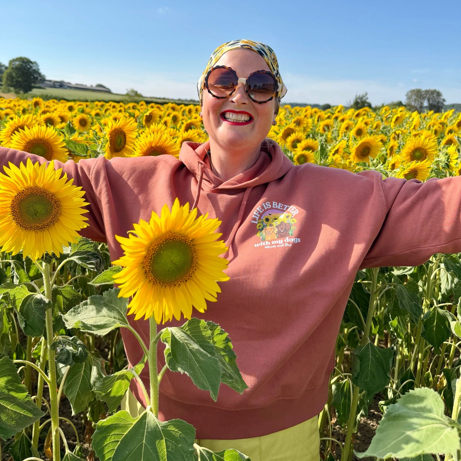 A woman wearing a brown hoody and sunglasses stands in a sunflower field with her arms outstretched, surrounded by bright yellow sunflowers under a clear blue sky.