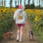 A woman with pink hair walks away in a sunflower field with two dogs on leashes. She is wearing a light gray hoodie with a large graphic on the back that reads 'Life is Better with my dogs' 