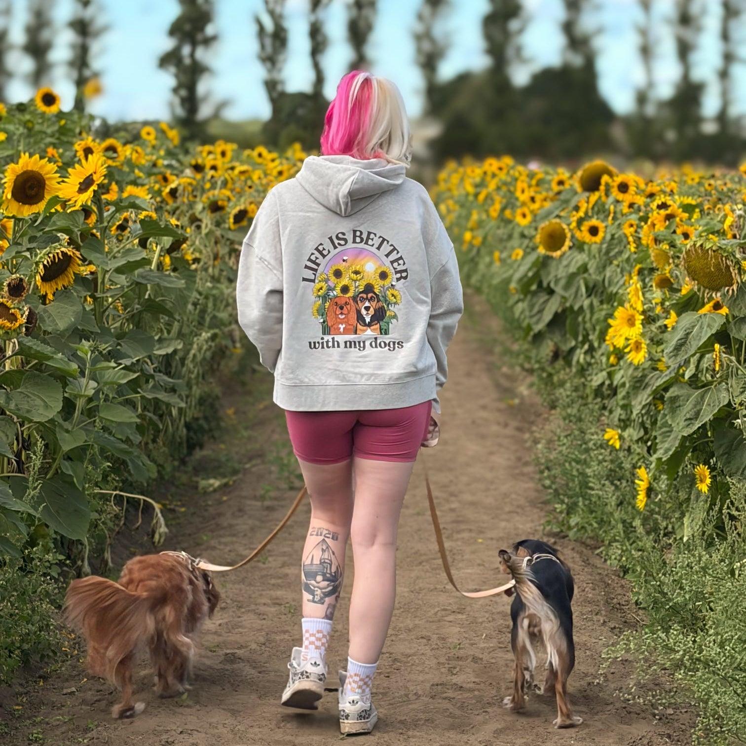 A woman with pink hair walks away in a sunflower field with two dogs on leashes. She is wearing a light gray hoodie with a large graphic on the back that reads 'Life is Better with my dogs' 