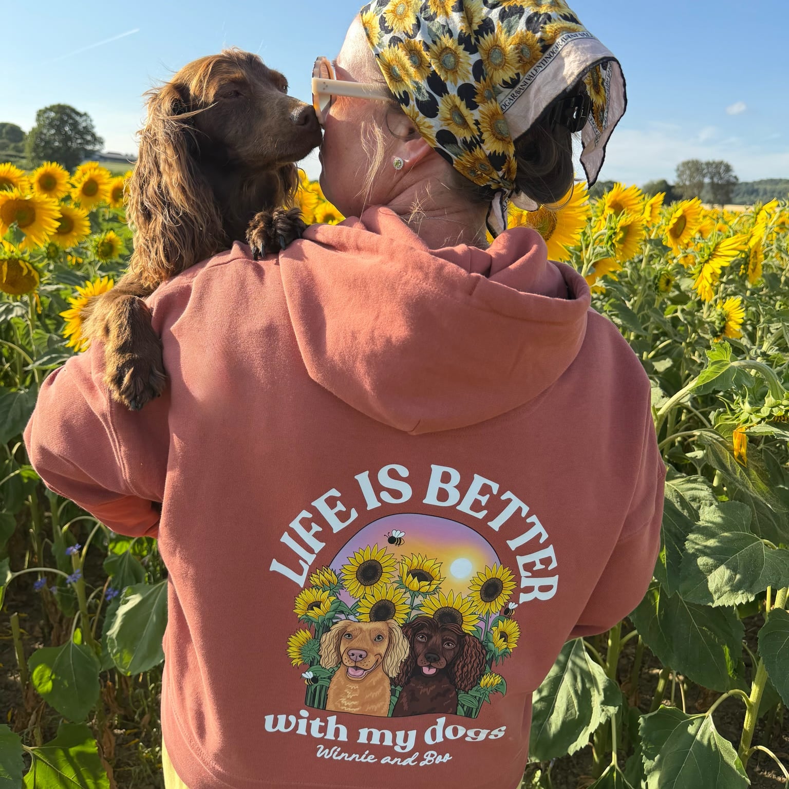 A person standing in a sunflower field wearing a brown hoody with a large graphic on the back that reads 'LIFE IS BETTER with my dogs' and shows a dog kissing the person's head. The person is also wearing a yellow and black patterned headscarf and yellow pants.
