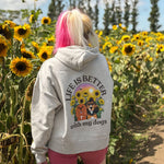 A person standing in a sunflower field, viewed from behind, wearing a light gray hoodie with a colorful graphic on the back that reads 'LIFE IS BETTER with my dogs' The sky is blue with white clouds in the background.