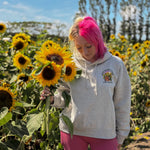 A woman with bright pink hair stands in a sunflower field wearing a light gray hoodie with a colorful sunflower graphic on the chest and pink shorts, holding a large sunflower.