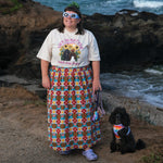 A woman stands on a rocky beach path wearing a white graphic t-shirt and a long, colorful geometric patterned maxi skirt, with a small black dog sitting beside her. The ocean waves and dark rocks are in the background.