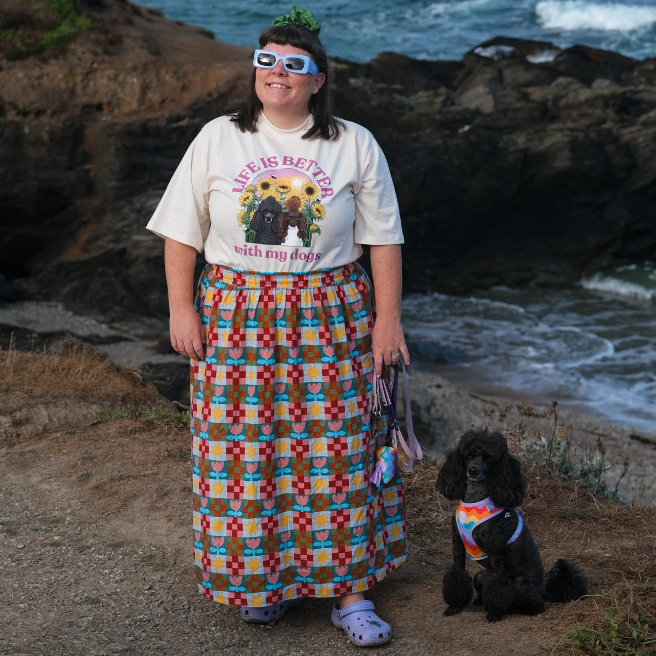 A woman stands on a rocky beach path wearing a white graphic t-shirt and a long, colorful geometric patterned maxi skirt, with a small black dog sitting beside her. The ocean waves and dark rocks are in the background.