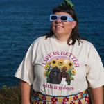 A person standing outdoors by the ocean wearing a cream graphic T-shirt with a dog print and the words 'LIFE IS BETTER with my dogs,' along with blue-framed sunglasses and a colorful patterned skirt.