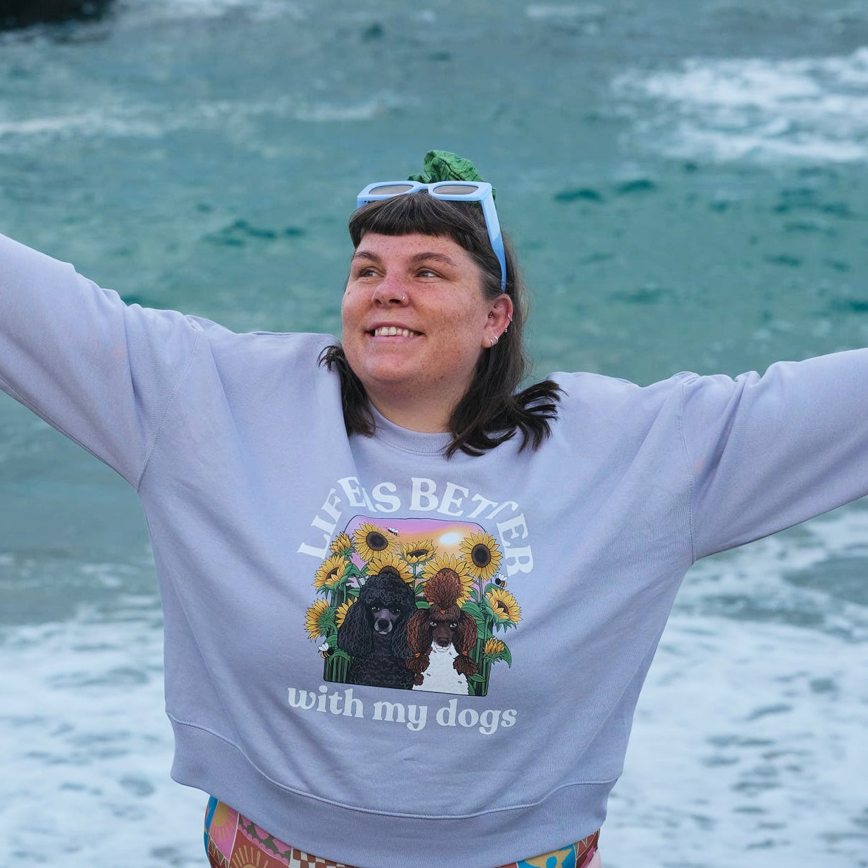 A woman stands on a beach with her arms outstretched, wearing a lavender sweatshirt with a graphic print that reads 'life is Better with my dogs' and a colorful patterned bikini. The ocean and rocks are visible in the background.