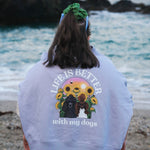 A person standing on a beach facing the ocean, wearing a light lavender sweatshirt with a large graphic on the back that reads 'LIFE IS BETTER with my dogs' and shows a dog and sunflowers, paired with colorful patterned bikini bottoms.