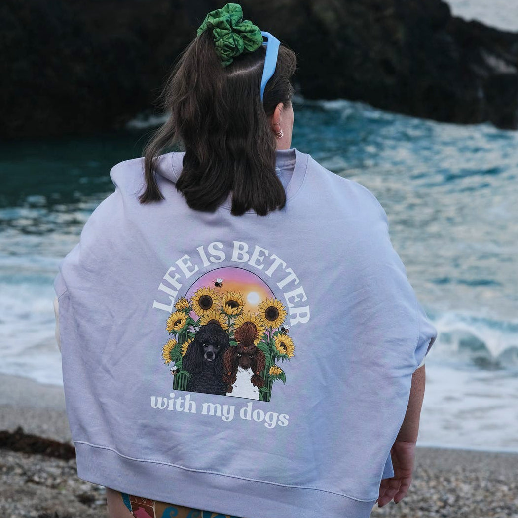 A woman standing on a rocky beach with her back to the camera, wearing a lavender hoody with a large graphic on the back that reads 'LIFE IS BETTER with my dogs' and shows a dog with sunflowers. A small black dog stands nearby on the sand.