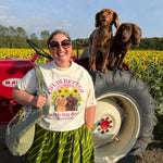 A woman wearing a graphic t-shirt and a green striped skirt stands in front of a red tractor with two brown dogs sitting on its tire, set against a background of a sunflower field.