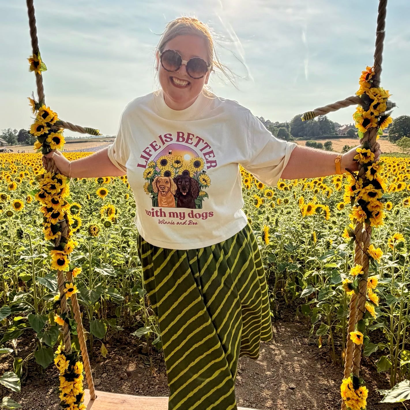 A woman stands on a rustic swing in a sunflower field, wearing a white graphic T-shirt that reads 'life is better with my dogs?' with a dog illustration, green and black striped pants, and sunglasses. The swing is decorated with yellow flower garlands.