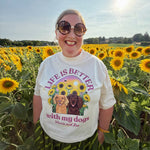 A woman wearing a white graphic t-shirt with a dog print and sunglasses stands in a sunflower field. The t-shirt features the text 'LIFE IS BETTER with my dogs' with illustrations of two dogs surrounded by sunflowers.