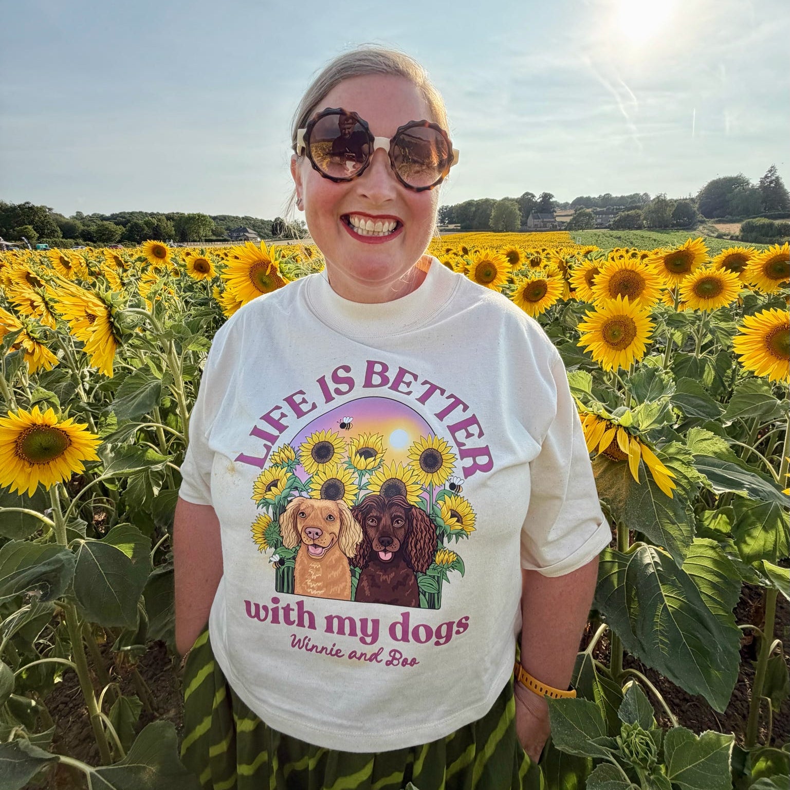 A woman wearing a white graphic t-shirt with a dog print and sunglasses stands in a sunflower field. The t-shirt features the text 'LIFE IS BETTER with my dogs' with illustrations of two dogs surrounded by sunflowers.