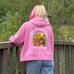 A person viewed from behind wearing a pink hoodie with a large graphic on the back that reads 'LIFE IS BETTER with my dogs' and shows two dogs surrounded by sunflowers. The person is standing on a wooden deck with green trees in the background.