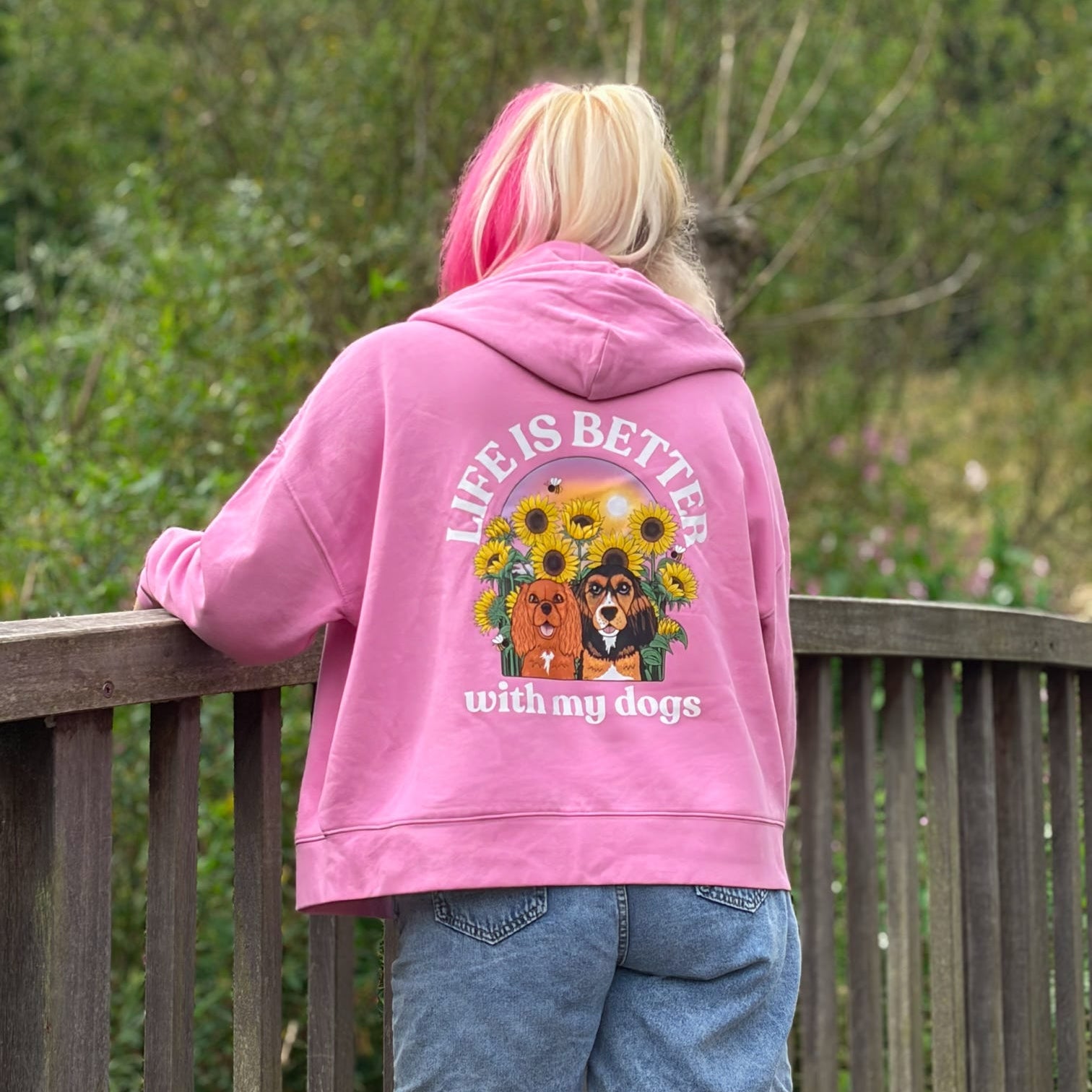 A person viewed from behind wearing a pink hoodie with a large graphic on the back that reads 'LIFE IS BETTER with my dogs' and shows two dogs surrounded by sunflowers. The person is standing on a wooden deck with green trees in the background.