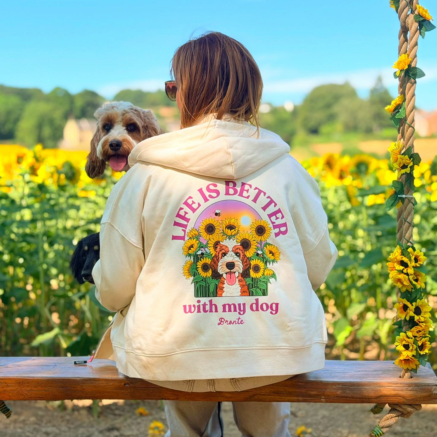A woman sits on a wooden swing in a sunflower field, viewed from behind, wearing a cream hoodie with a large graphic on the back that reads 'LIFE IS BETTER with my dog' and shows a dog and sunflowers. A real dog is visible beside her.
