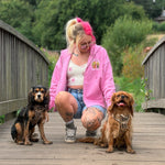 A woman crouches on a wooden bridge outdoors with two dogs. She is wearing a bright pink zip-up hoodie, denim shorts, and sneakers.