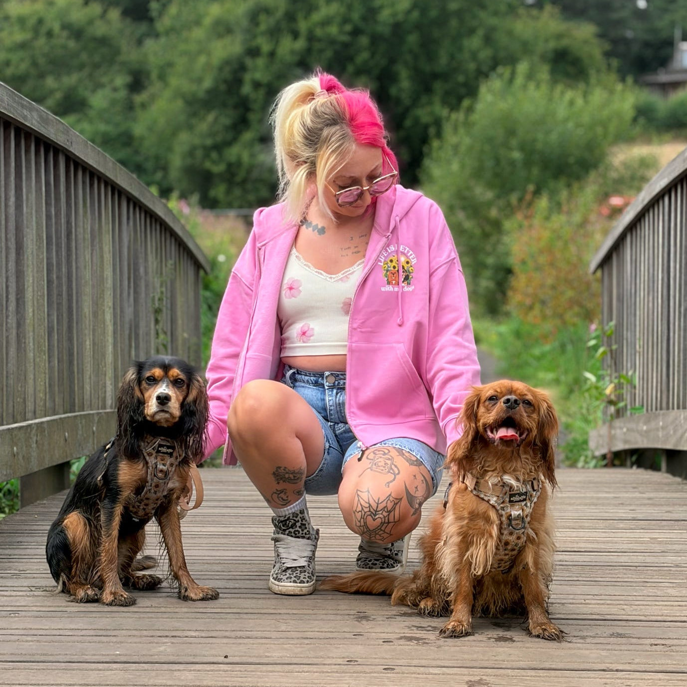 A woman crouches on a wooden bridge outdoors with two dogs. She is wearing a bright pink zip-up hoodie, denim shorts, and sneakers.
