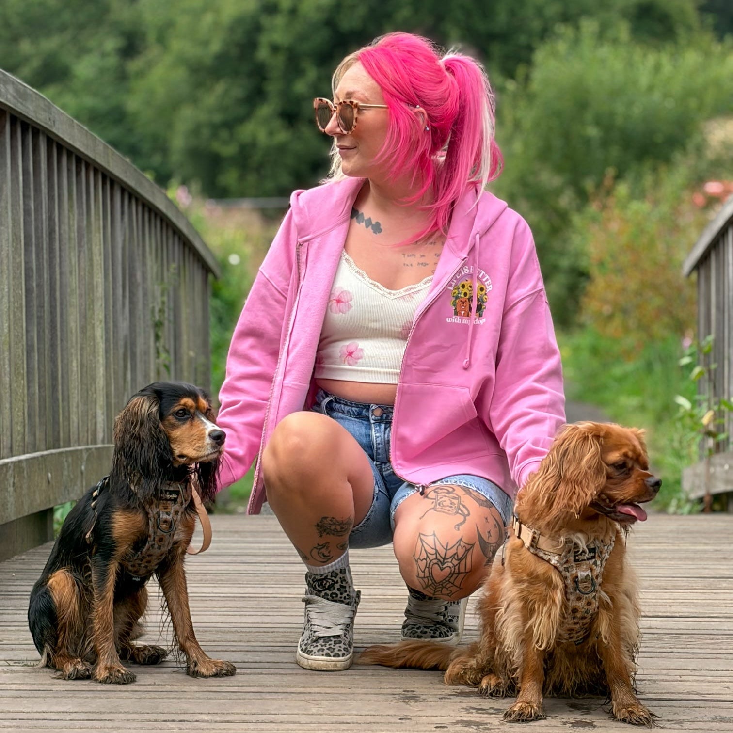 A woman with bright pink hair kneels on a wooden boardwalk outdoors, wearing a pink zip-up hoodie, denim shorts, and patterned socks, while sitting between two dogs. Trees and a wooden bridge are visible in the background.