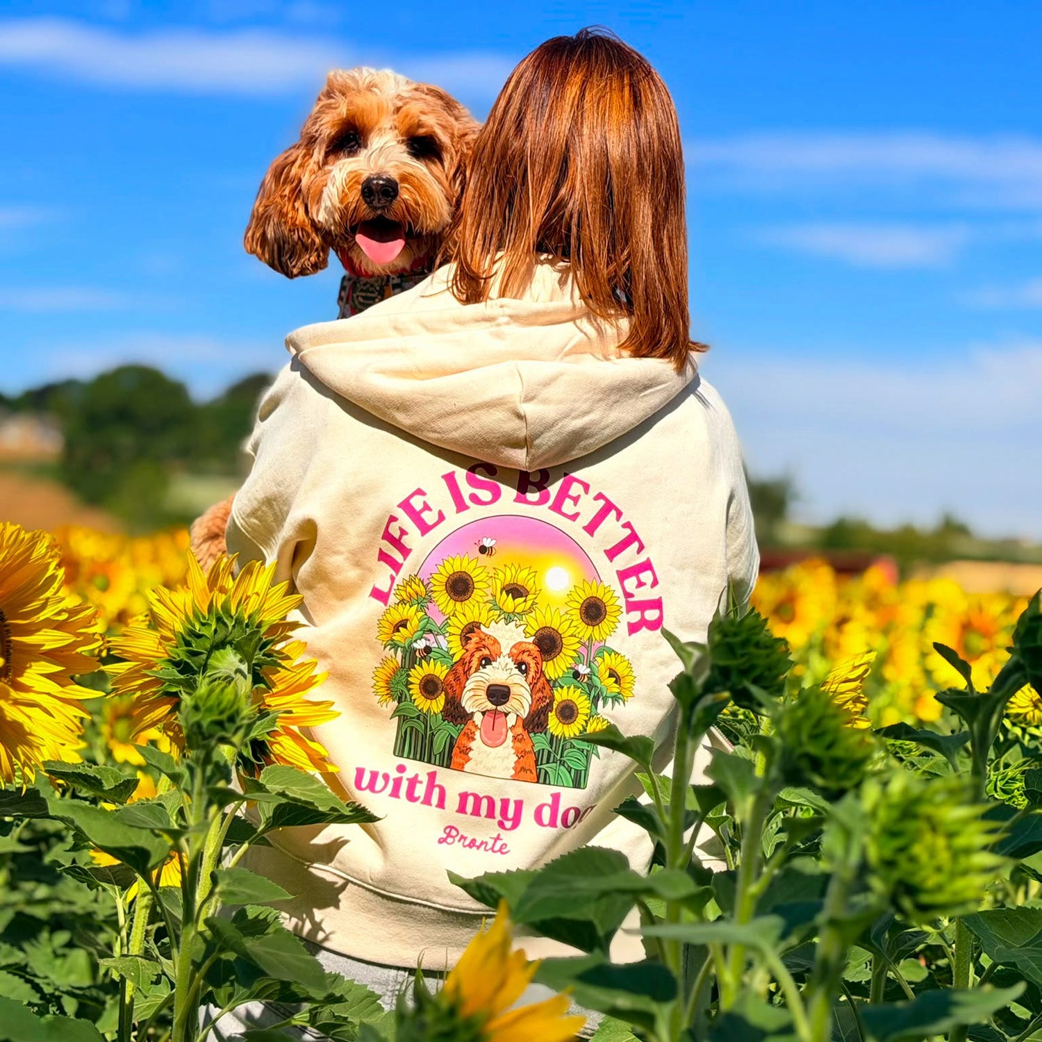 A woman and her brown dog are seen from behind in a sunflower field, wearing a cream hoody with a colorful graphic that reads 'Life is better with my dog'.