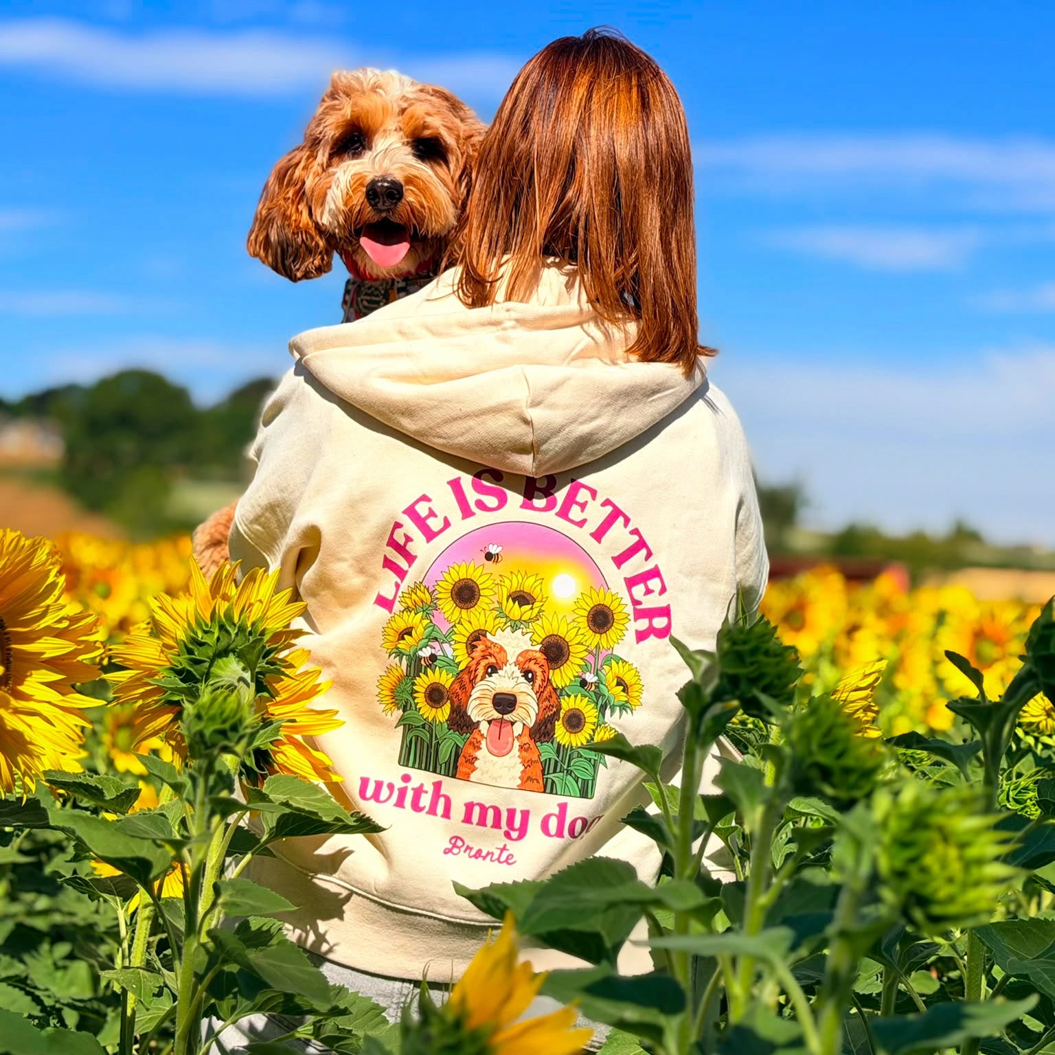 A woman and her brown dog are seen from behind in a sunflower field, wearing a cream hoody with a colorful graphic that reads 'Life is better with my dog'.