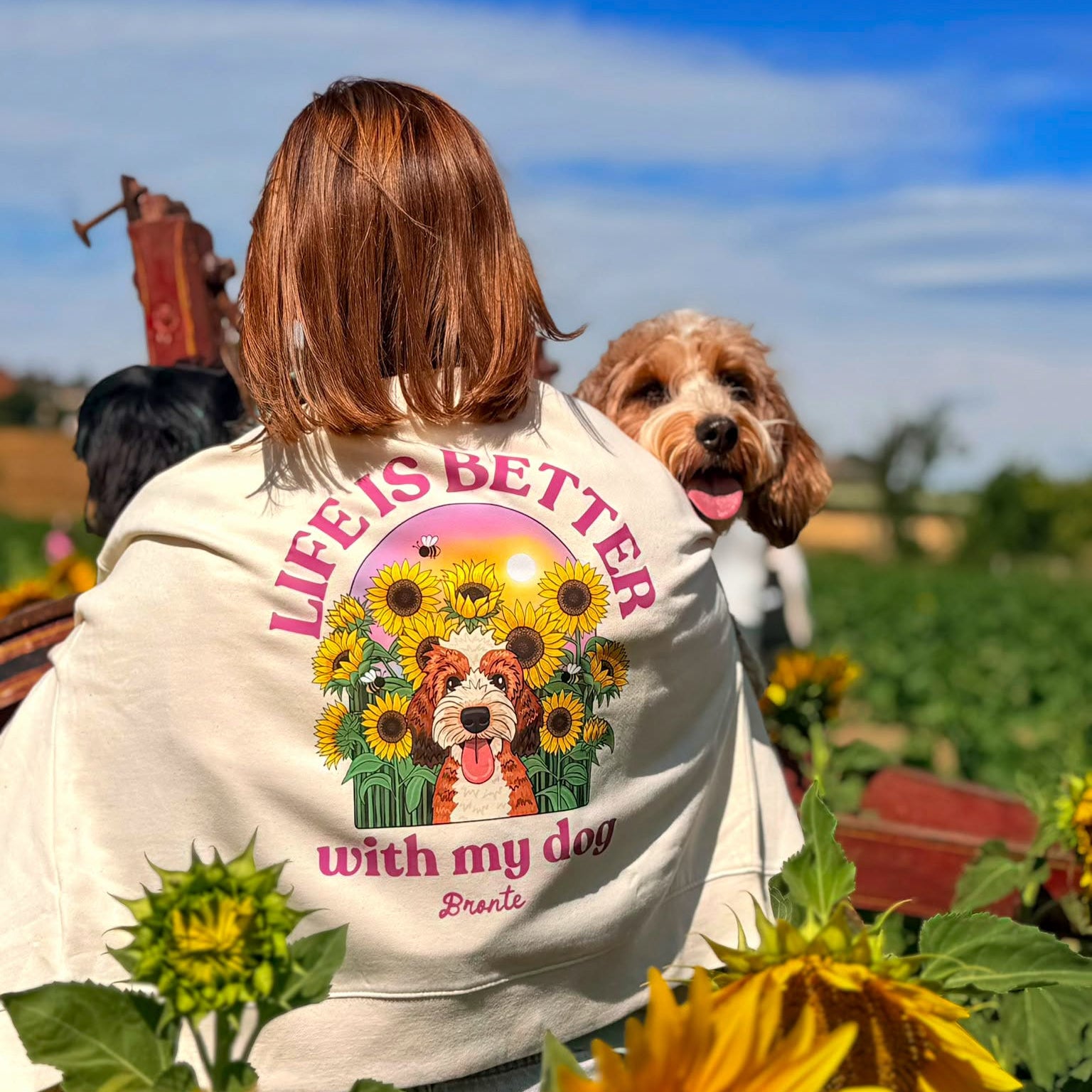 A woman seen from behind wearing a cream-colored hoody with a colorful graphic of a dog surrounded by sunflowers and the text 'LIFE IS BETTER with my dog'. A brown and white dog sits on her shoulder in an outdoor field of sunflowers under a blue sky.