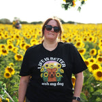 A woman wearing a black graphic t-shirt stands in a sunflower field. The t-shirt features a dog illustration amongst some sunflowers with the text 'LIFE IS BETTER with my dog'.