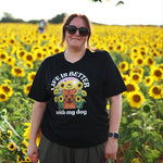 A woman wearing a black graphic t-shirt and sunglasses stands in a sunflower field. The t-shirt features a dog illustration amongst some sunflowers and the text 'LIFE IS BETTER with my dog'.