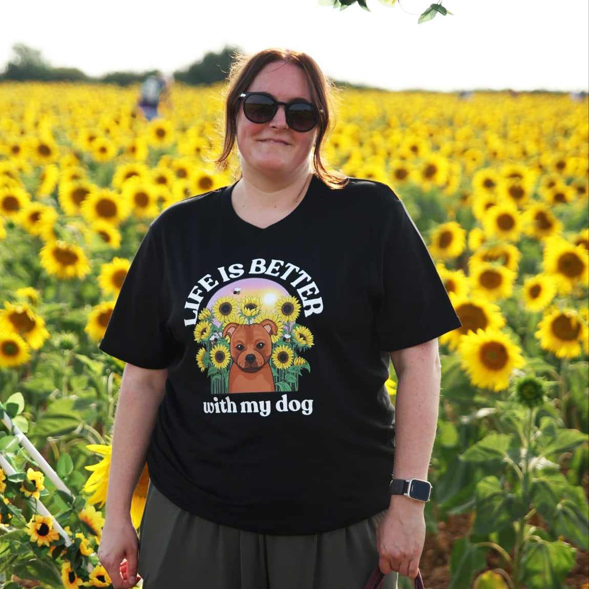 A woman wearing a black graphic t-shirt and sunglasses stands in a sunflower field. The t-shirt features a dog illustration amongst some sunflowers and the text 'LIFE IS BETTER with my dog'.