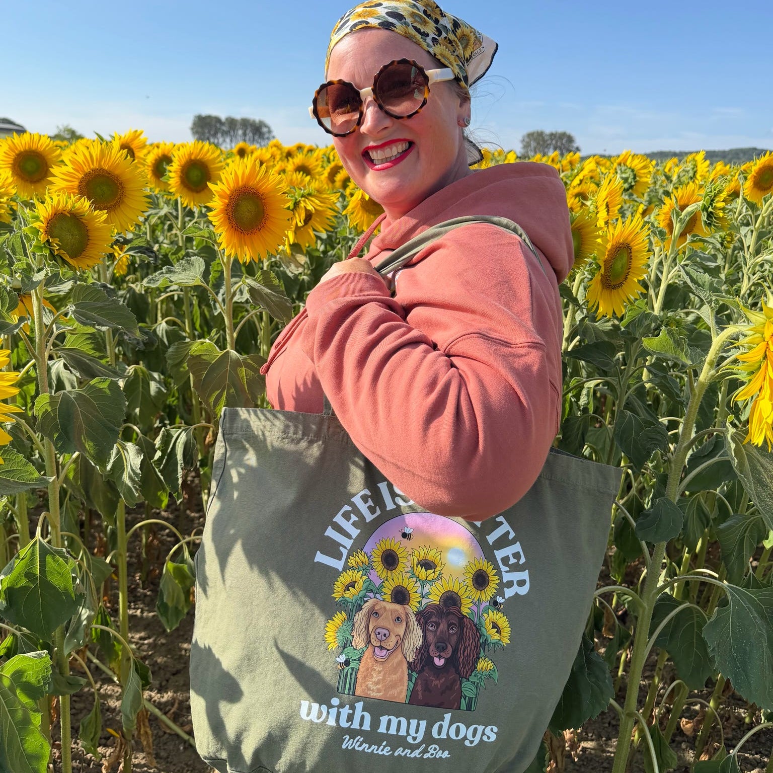 A woman standing in a sunflower field wearing an ornage hoodie, sunglasses, and a patterned headscarf, carrying a large olive green tote bag printed with 'life is better with my dogs' and an illustration of two dogs with sunflowers. The background shows a field of sunflowers under a blue sky.