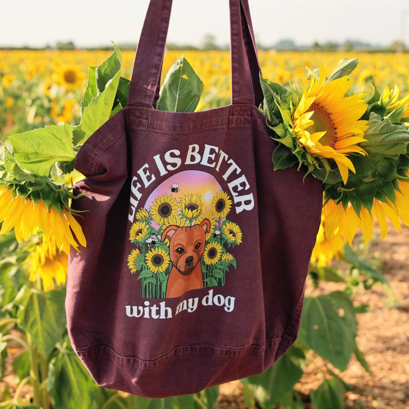 A person's hand holds a dark purple canvas tote bag printed with 'LIFE IS BETTER with my dog' and a dog surrounded by sunflowers. The bag is framed by real sunflowers in a sunny sunflower field.