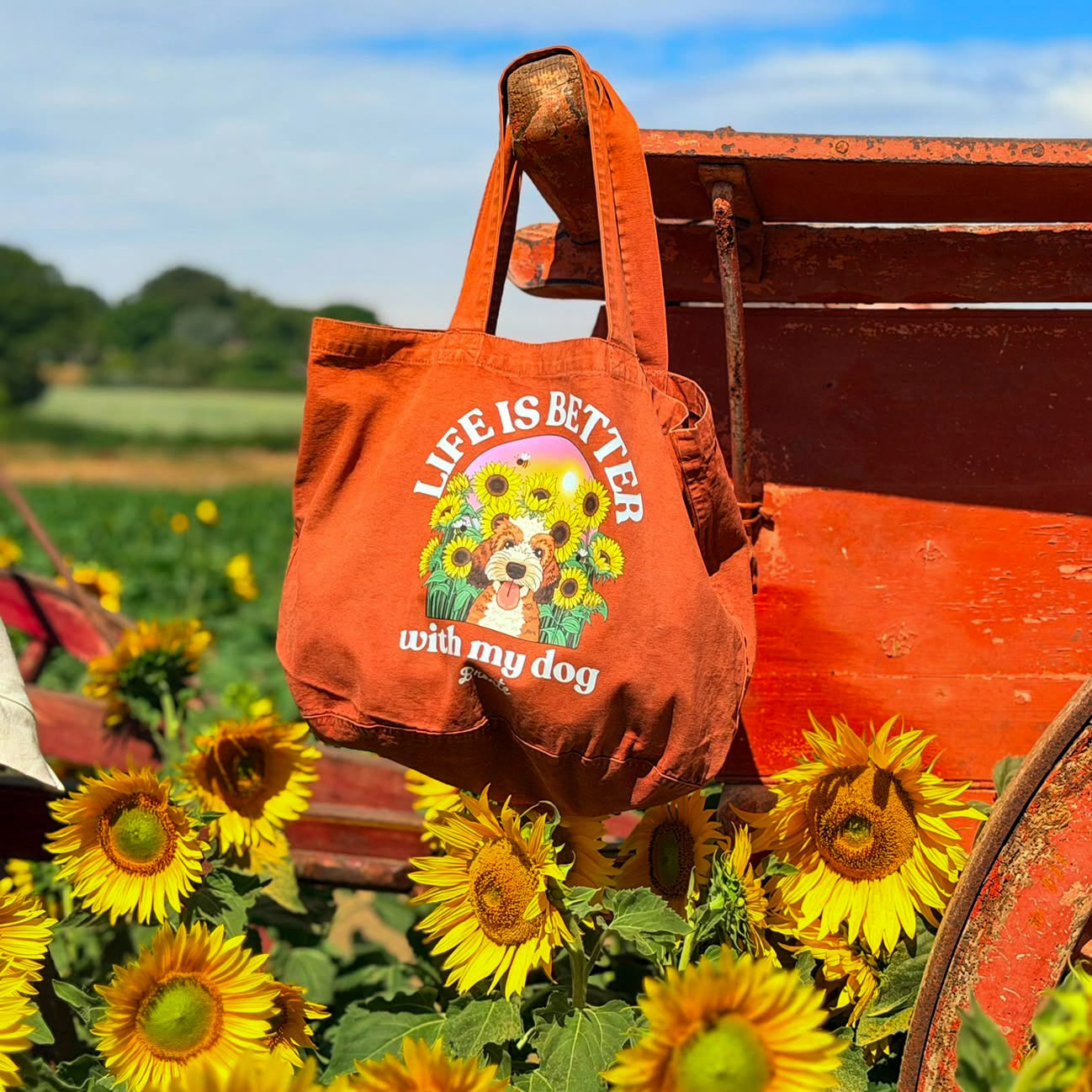 An orange tote bag with a dog graphic and the text 'LIFE IS BETTER with my dog' hanging from a weathered red wagon in a sunflower field under a blue sky.
