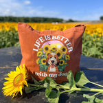An orange-brown canvas tote bag with a colorful graphic of a dog surrounded by sunflowers and the text 'LIFE IS BETTER with my dog' sits on a dark table outdoors in front of a sunflower field under a blue sky. A real sunflower rests in the foreground.