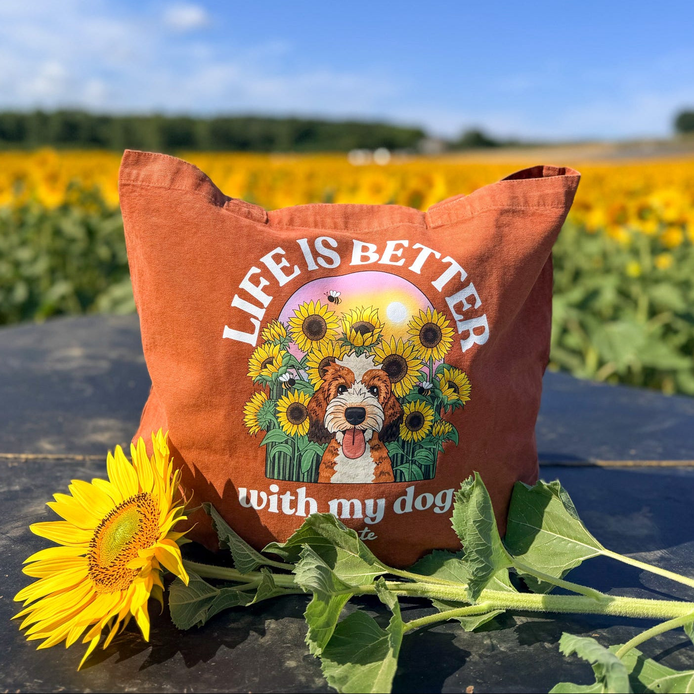 An orange-brown canvas tote bag with a colorful graphic of a dog surrounded by sunflowers and the text 'LIFE IS BETTER with my dog' sits on a dark table outdoors in front of a sunflower field under a blue sky. A real sunflower rests in the foreground.