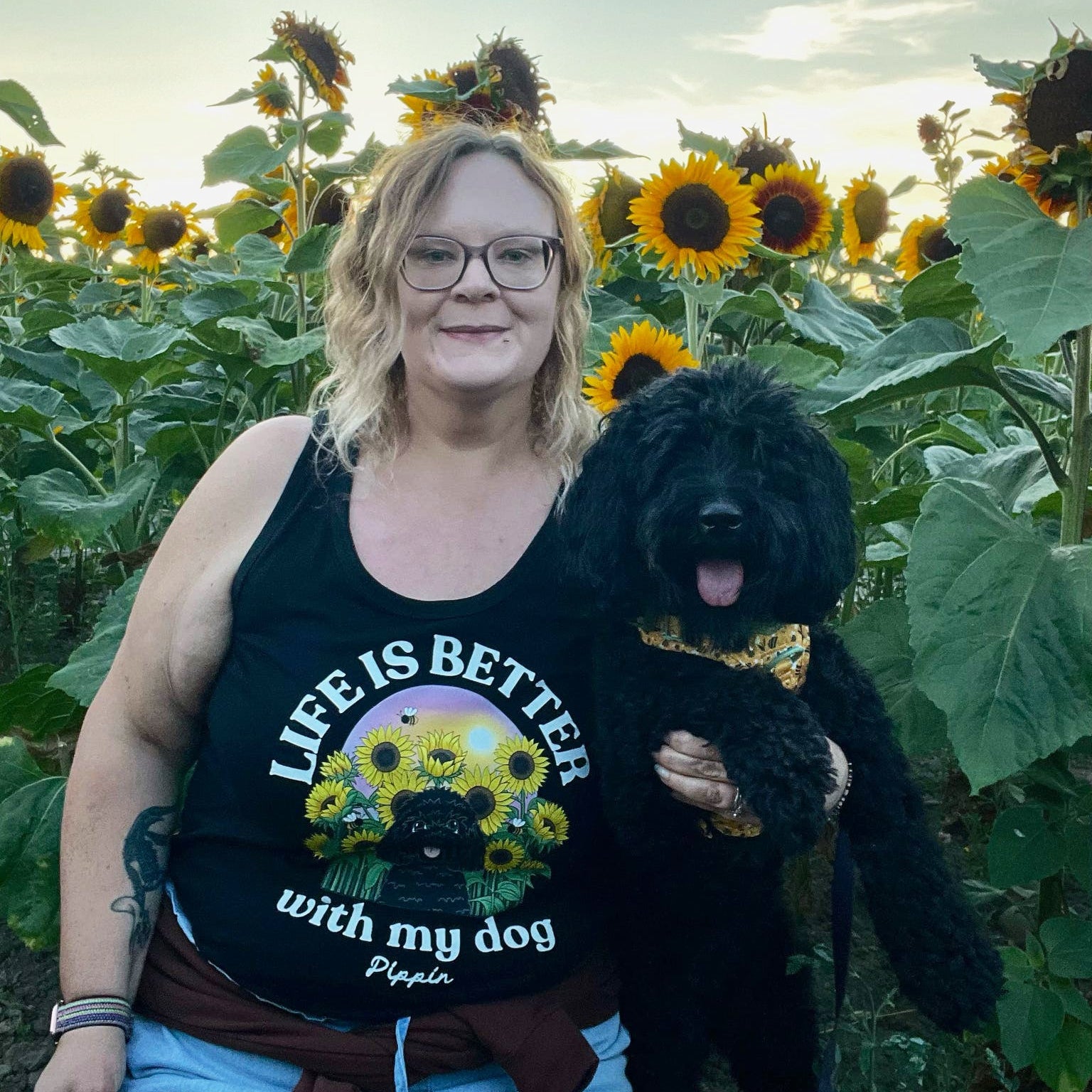 A woman wearing a black graphic vest top with a dog and sunflower design, standing in a sunflower field and holding a black dog.