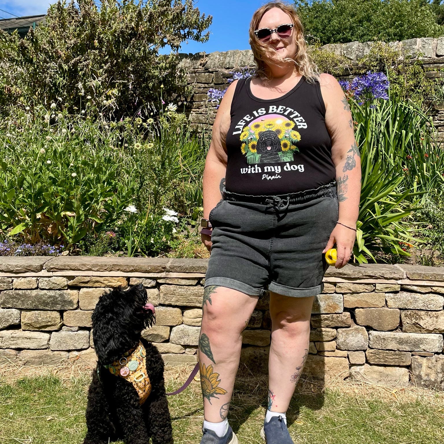 A woman stands outdoors on a grassy lawn in front of a stone wall and garden plants, wearing a black graphic vest top that reads 'Life is better with my dog' and dark shorts. A small black dog sits beside her wearing a patterned bandana.