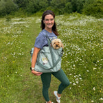 Woman holding a dog in a carrier while standing in a field with white flowers and green grass.