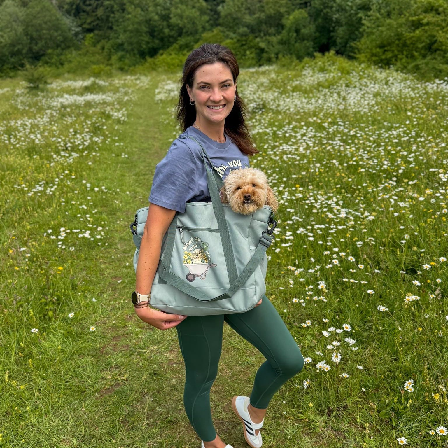 Woman holding a dog in a carrier while standing in a field with white flowers and green grass.