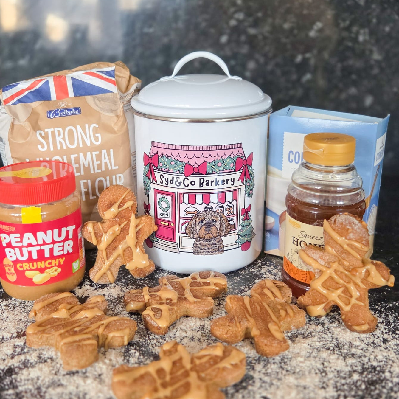 Dog treats on a dark surface with flour, peanut butter, and a jar of honey.