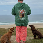 Person wearing a green hoodie with a dog-themed design, standing on a beach with two dogs.