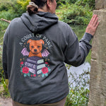 A woman viewed from behind wearing a dark gray hoodie with a large graphic on the back featuring a dog, books, roses, and the text 'A COURT OF PEACE'. She is standing outdoors next to a stone pillar with a lush green forest and river in the background.