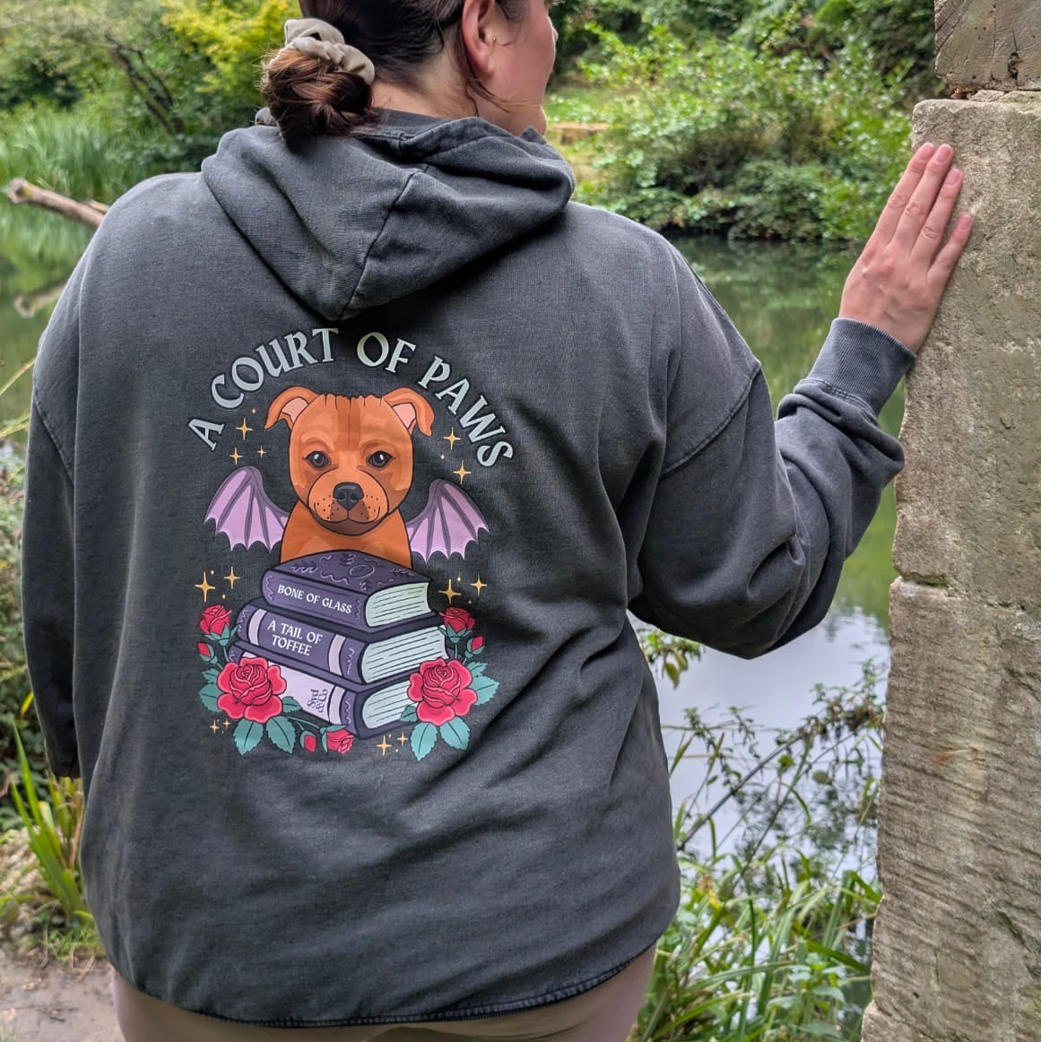 A woman viewed from behind wearing a dark gray hoodie with a large graphic on the back featuring a dog, books, roses, and the text 'A COURT OF PEACE'. She is standing outdoors next to a stone pillar with a lush green forest and river in the background.