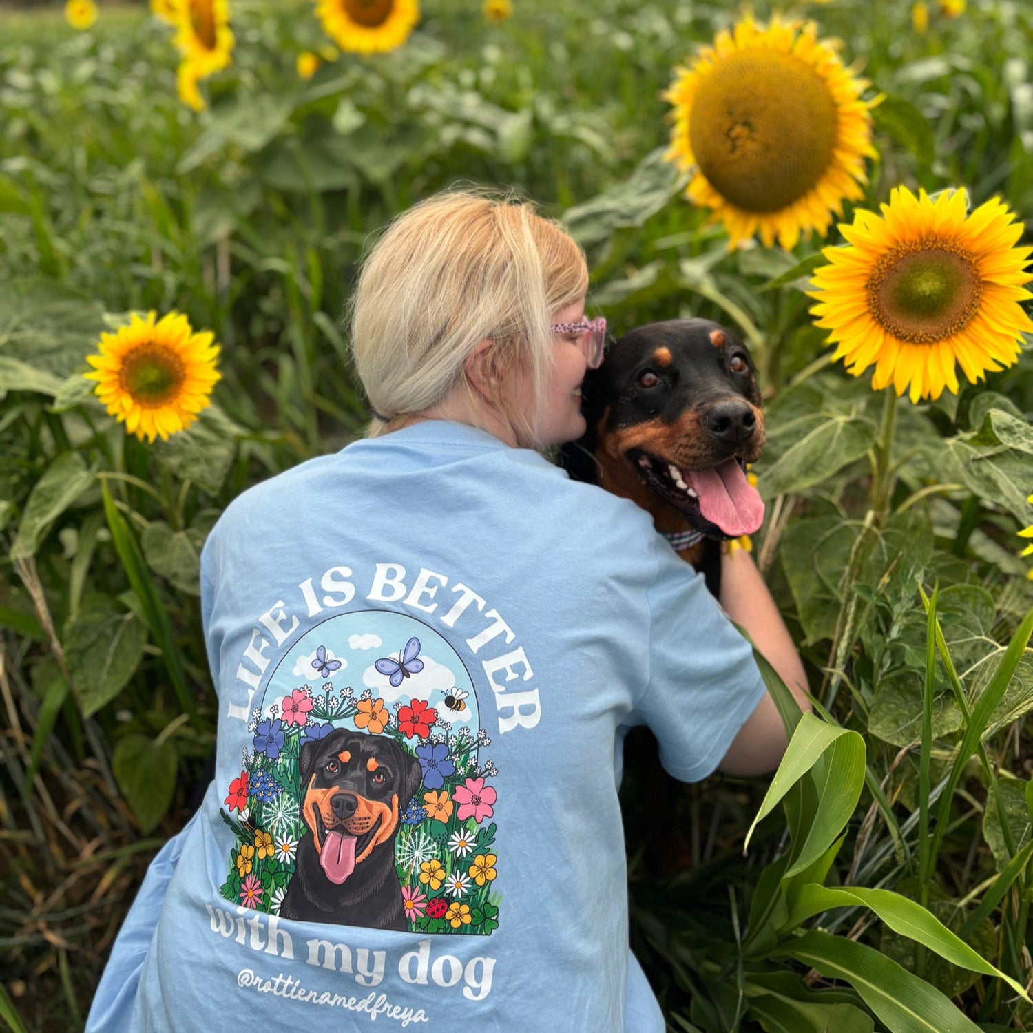Person in a sunflower field holding a dog, wearing a shirt with a dog-themed design.