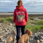Person wearing a red hoodie with a dog graphic and text, standing on a rocky beach with two dogs.