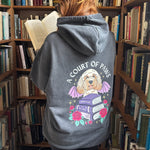 A person standing in a book-filled library aisle wearing a dark gray hoodie with a colorful graphic on the back of a dog sitting on books under the text 'A COURT OF PAWS'.
