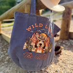 Blue tote bag with 'Wild Like The West' design and cowboy hat on wooden surface